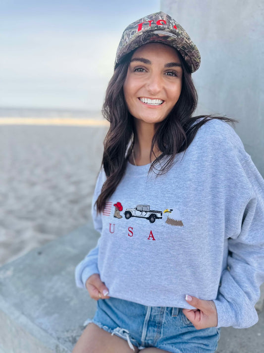 Woman wearing a light blue sweatshirt with an eagle, cowboy boots, truck and usa flag graphic design, sitting on a beach.