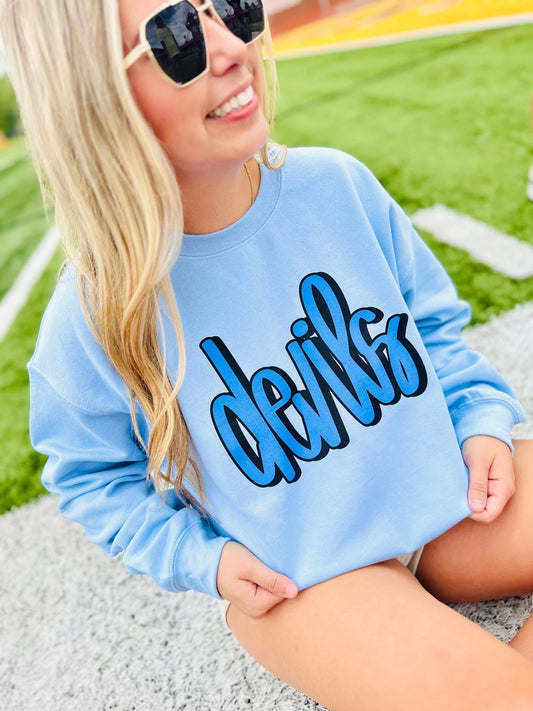 Person wearing a light blue sweatshirt with 'devils' printed on it, sitting on a grassy field.
