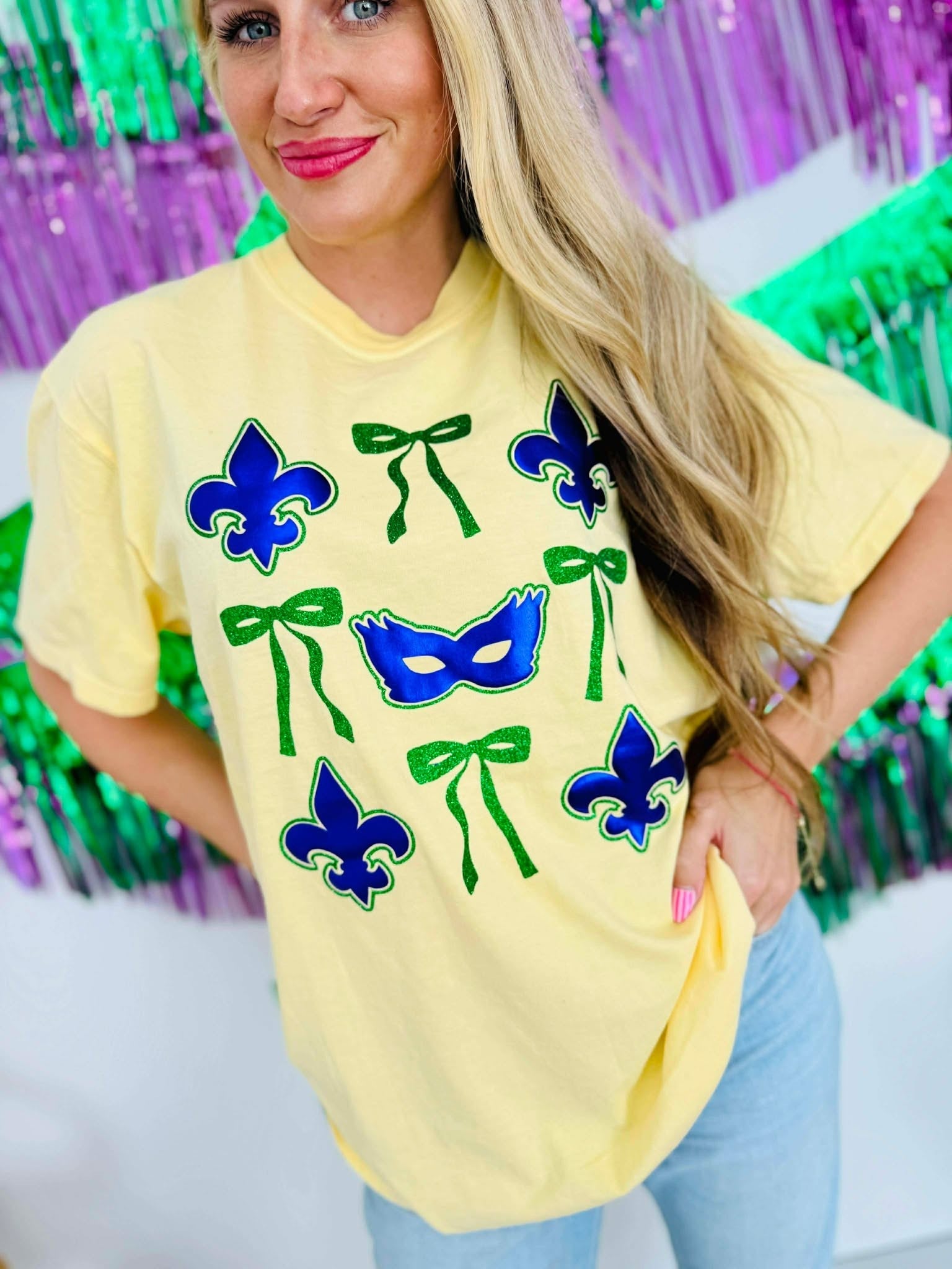 Woman wearing a yellow t-shirt with purple masks and green ribbons in front of a colorful background for Mardi Gras