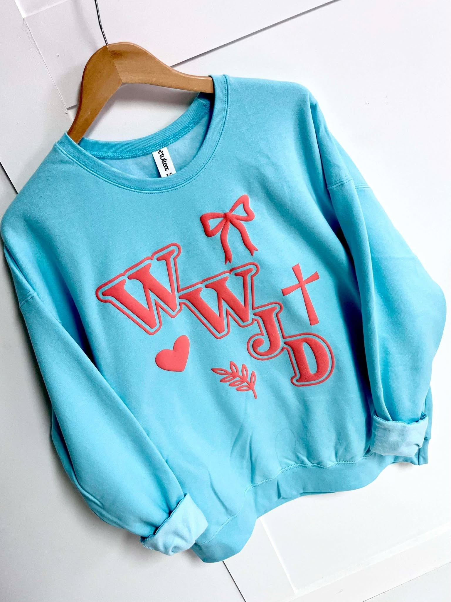 Light blue sweatshirt with coral puff text WWJD and cross, bow, heart leaf graphics on a white tiled floor