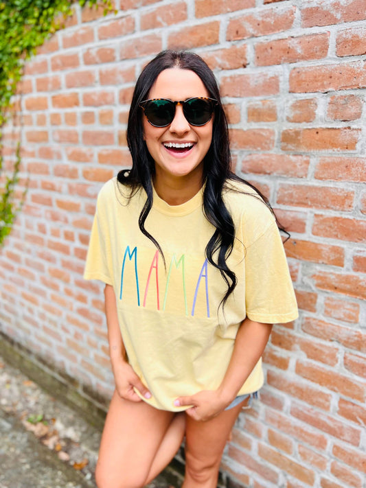 A person wearing a yellow graphic tee with the text 'MAMA' embroidered on the front, standing with one hand against a brick wall.