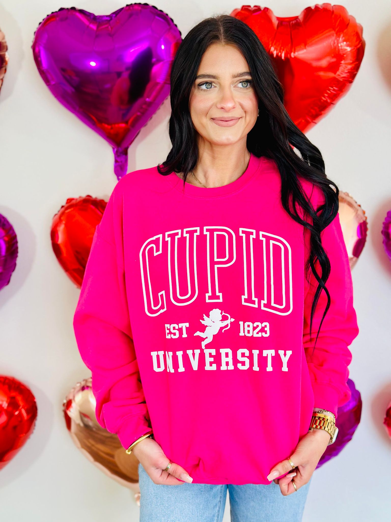 A person wearing a pink sweatshirt with the text 'CUPID UNSUCCESSFUL SINCE 1823' and a graphic of a cupid, surrounded by heart-shaped balloons.