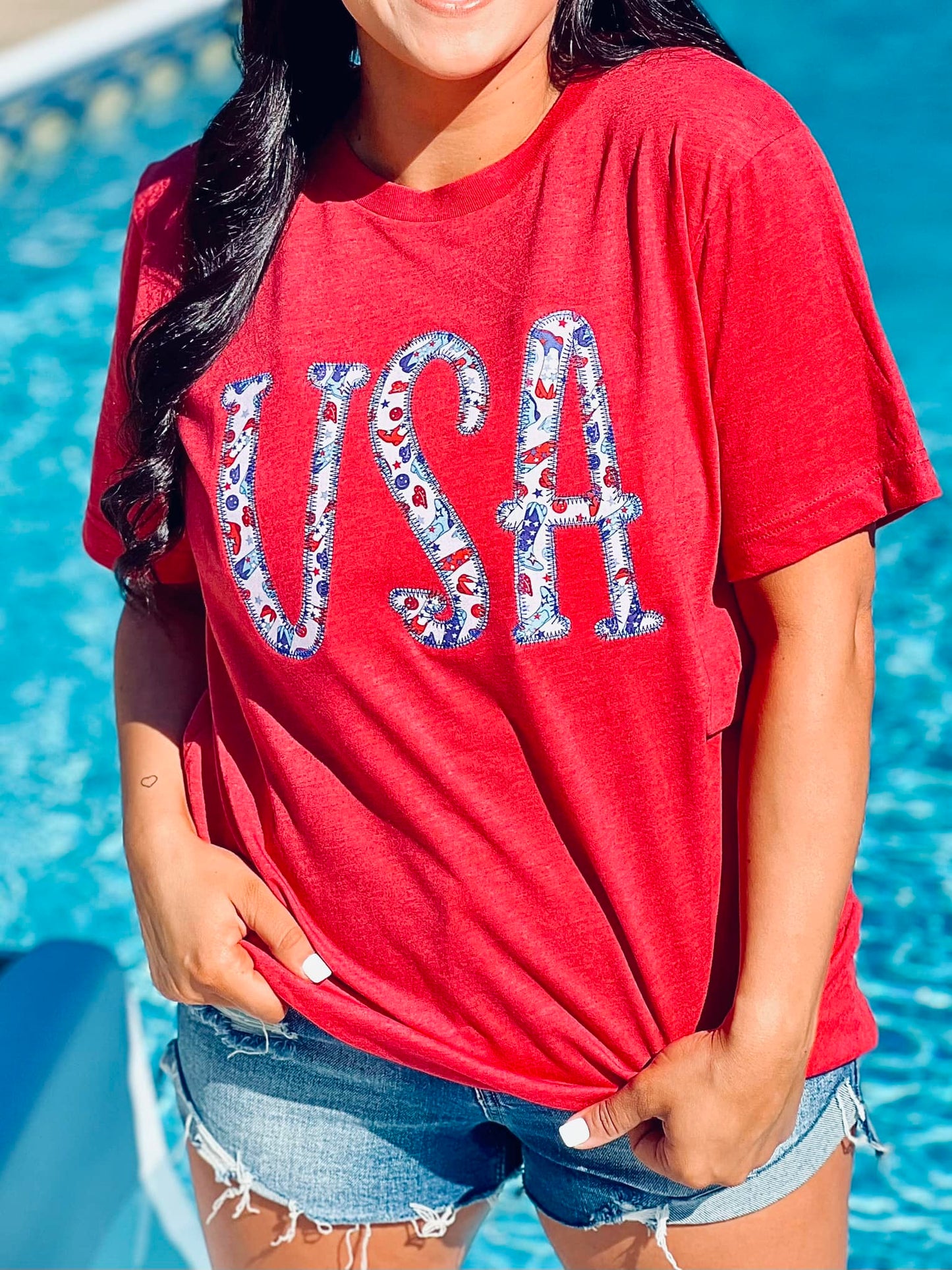 A person wearing a red t-shirt with the text 'USA' and an embroidered design resembling a flag on it, standing by a pool.
