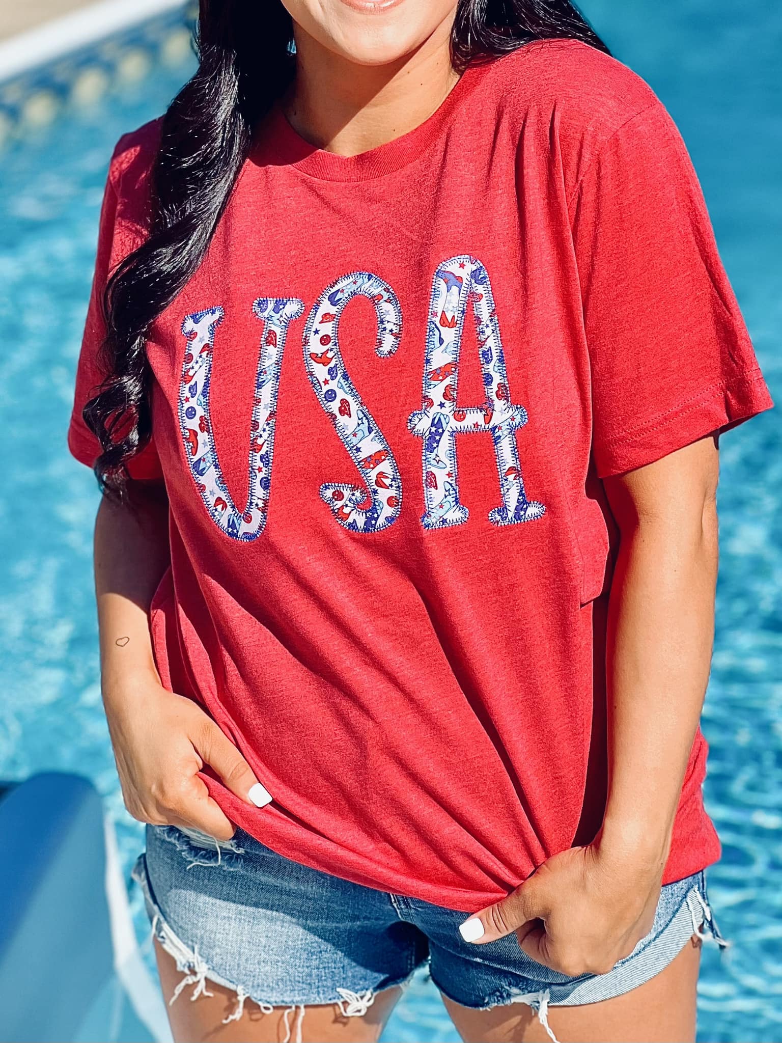 A person wearing a red t-shirt with the text 'USA' and an embroidered design resembling a flag on it, standing by a pool.