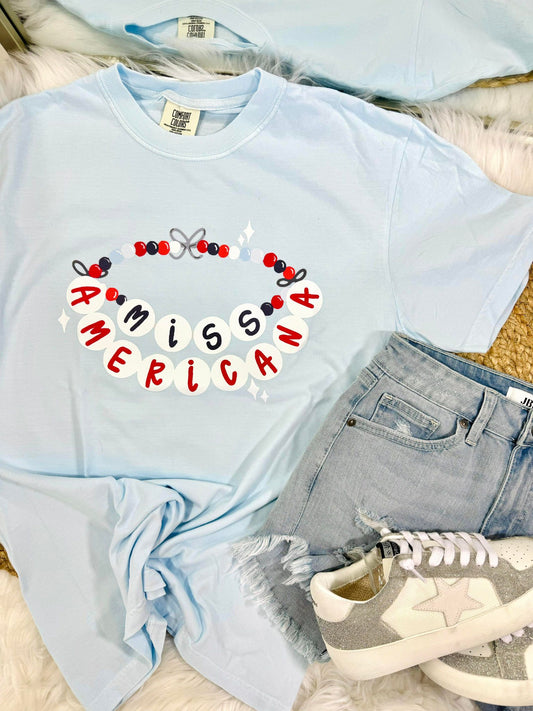 A light blue t-shirt with the text 'MISS AMERICA' and decorative friendship bracelet elements printed across the front, displayed flat with denim shorts and white sneakers.