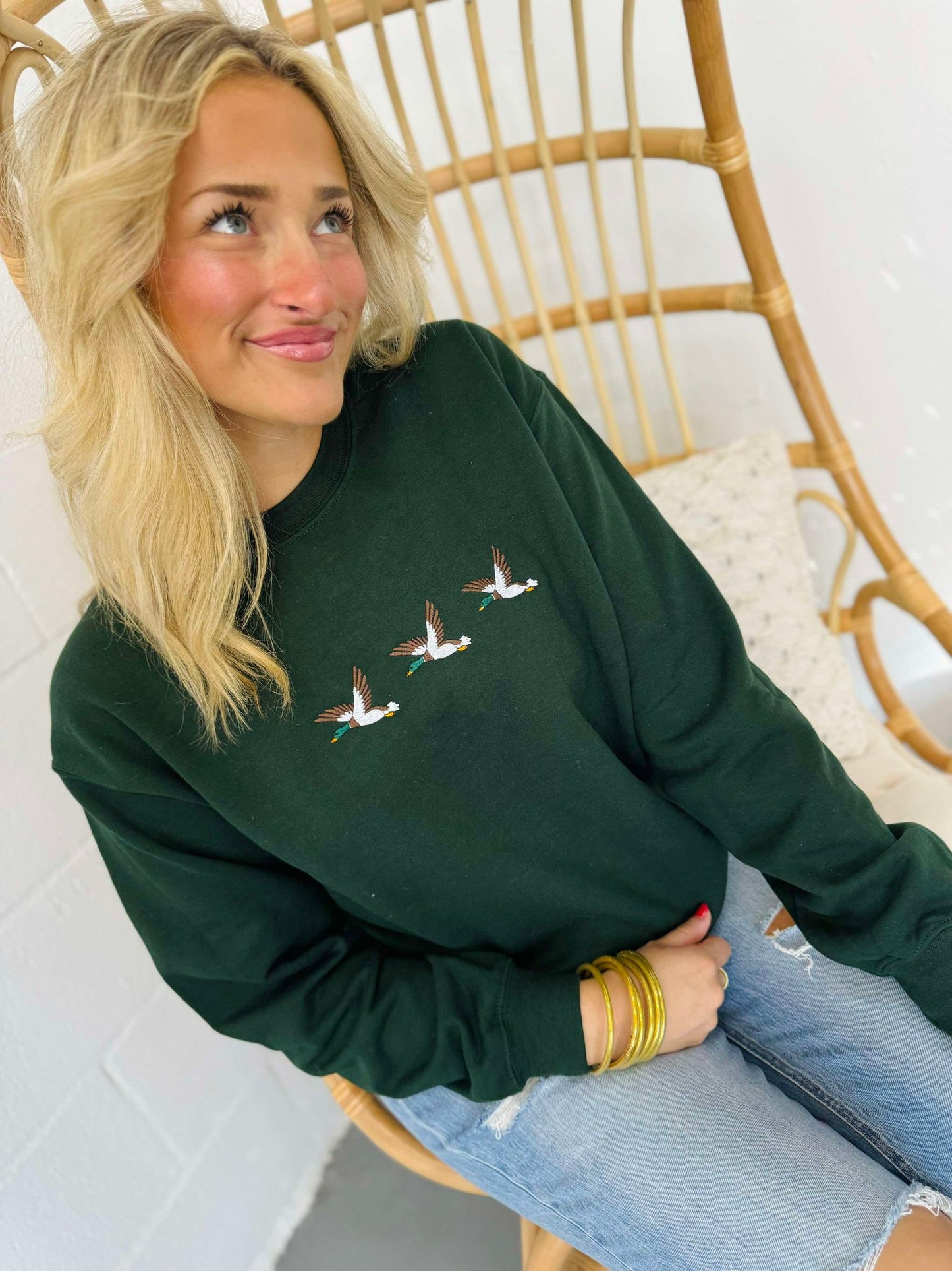 A woman sitting on a wooden chair wearing a green embroidered sweatshirt with a trio of mallard ducks on the front.