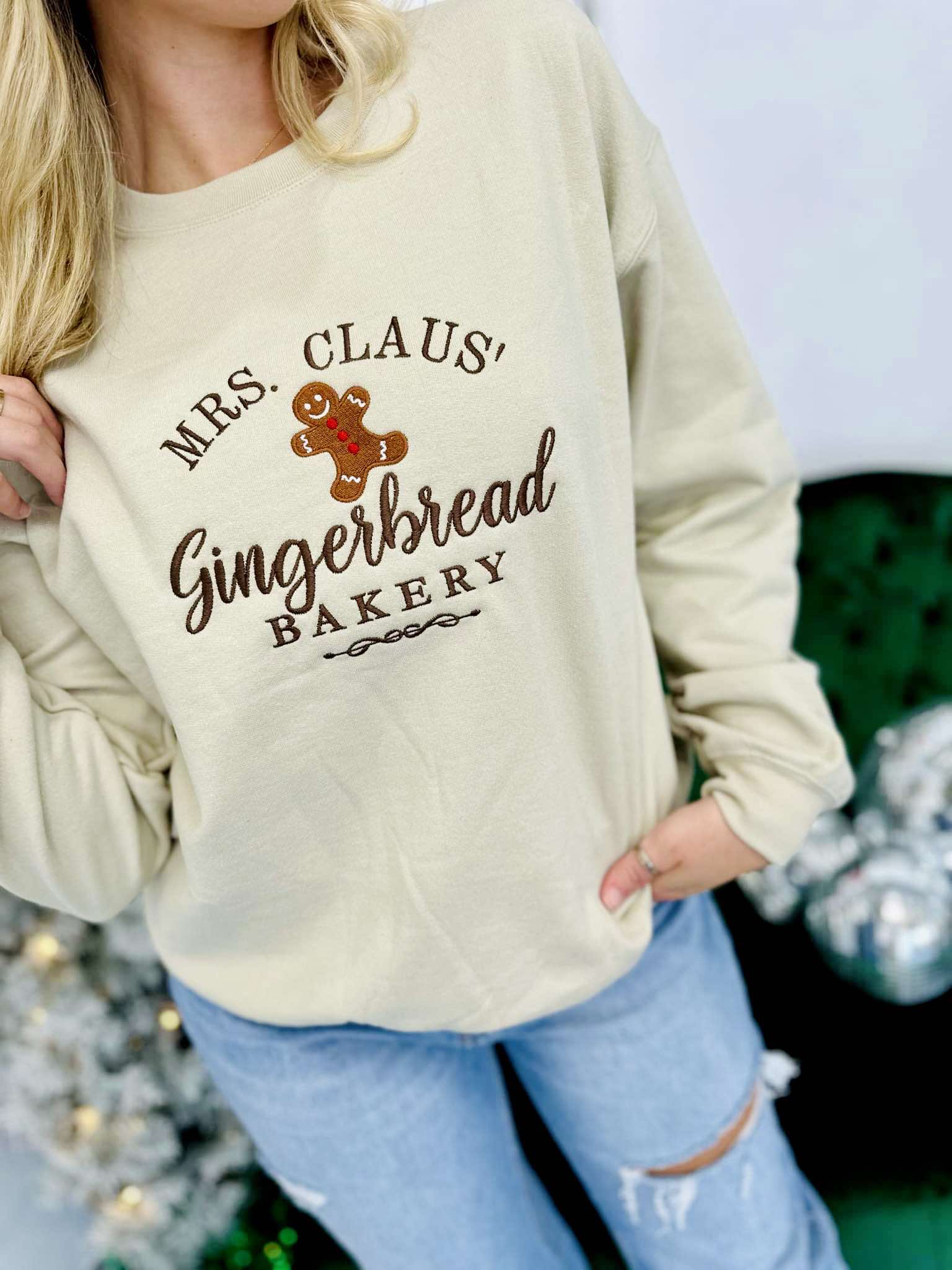 A woman wearing a beige embroidered sweatshirt with 'MRS. CLAUS' GINGERBREAD BAKERY' text and a cute gingerbread man graphic.