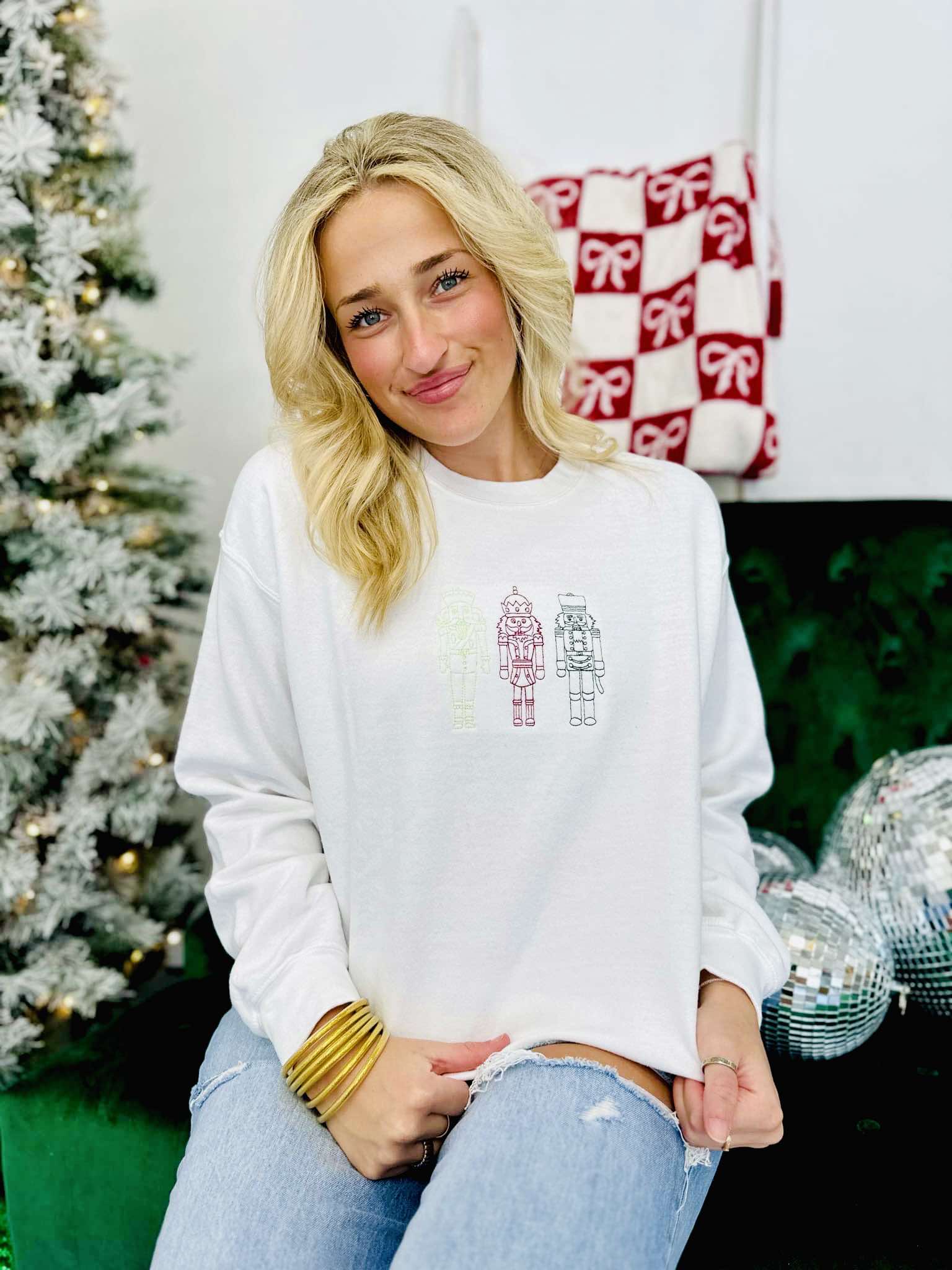 A woman sitting and wearing a white embroidered sweatshirt with the text 'Christmas Nutcracker Trio' on it, in front of a blurred Christmas tree and gifts in the background.