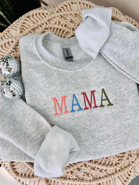 A gray sweatshirt with 'Mama' embroidered in red and green colors on the front, displayed in a woven basket with decorative balls and a tag.