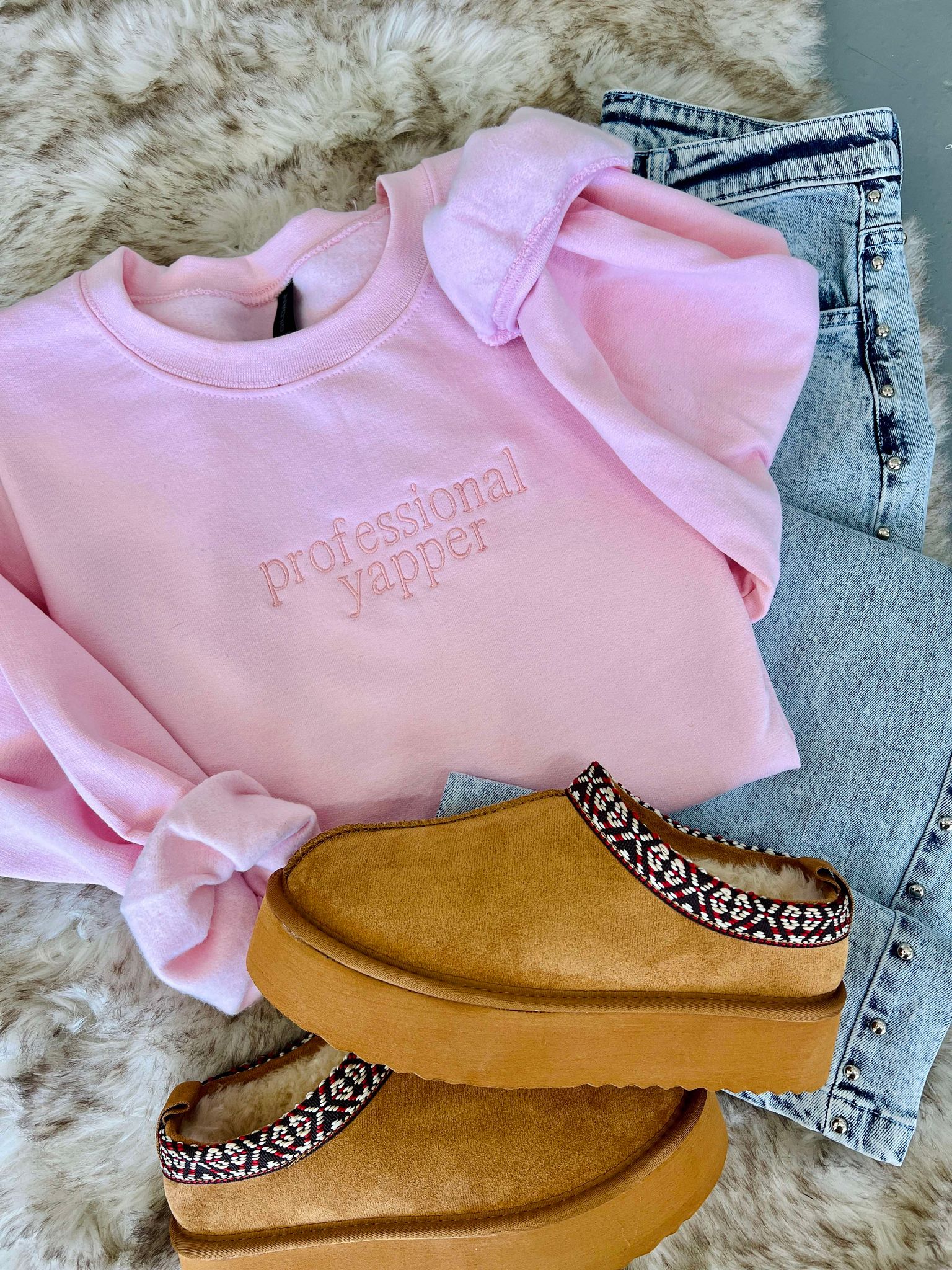 A light pink embroidered sweatshirt with the text 'PROFESSIONAL YAPPER' on the front, placed on a white furry surface, with a pair of blue jeans and a pair of tan casual shoes with embellished sole patterns on a matching white furry surface.