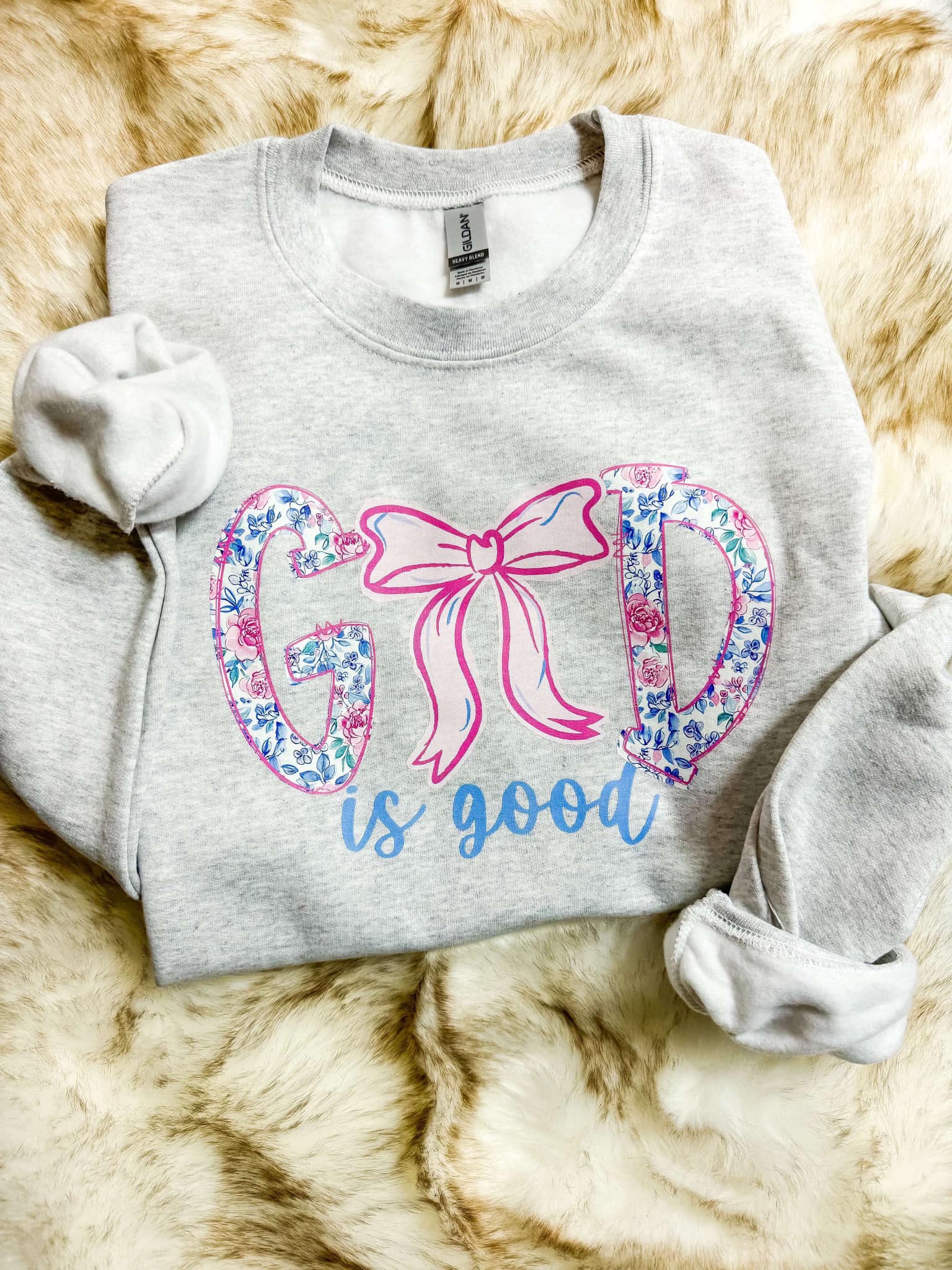 Ash grey sweatshirt with the phrase 'God is good' printed in a floral ribbon style in pink and blue.