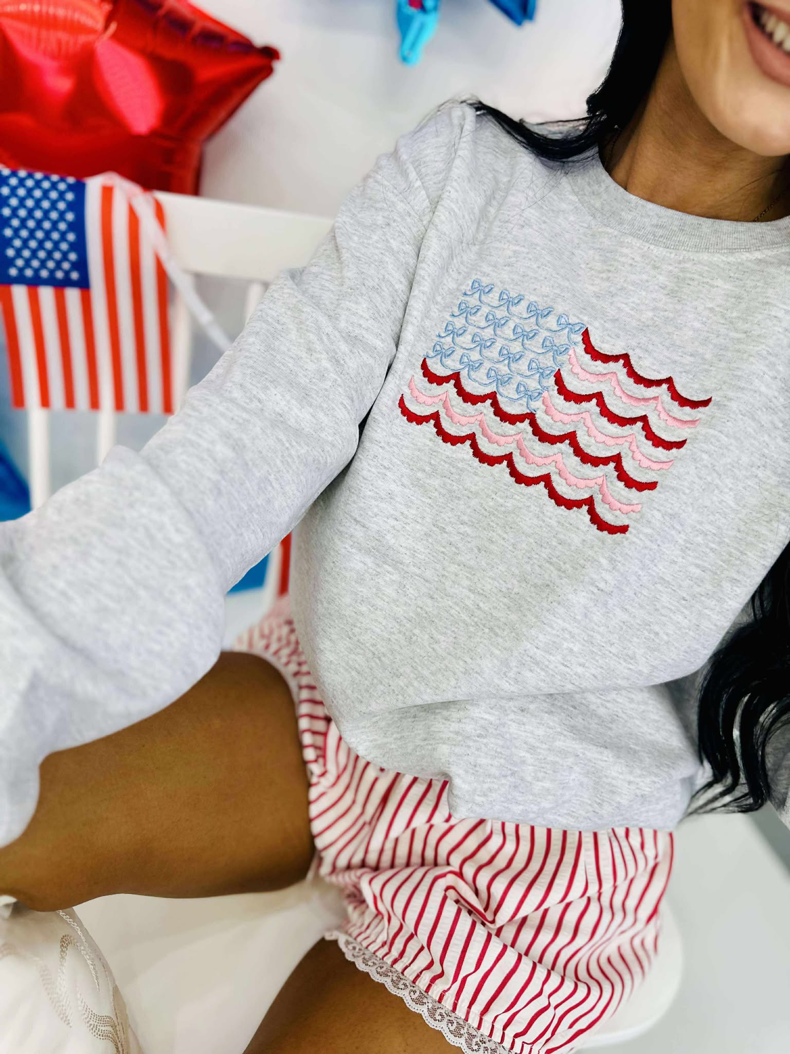 A sweatshirt with an embroidered American flag design in red and blue on a gray background, worn by a person seated in front of a small table decorated with American flags and related merchandise.
