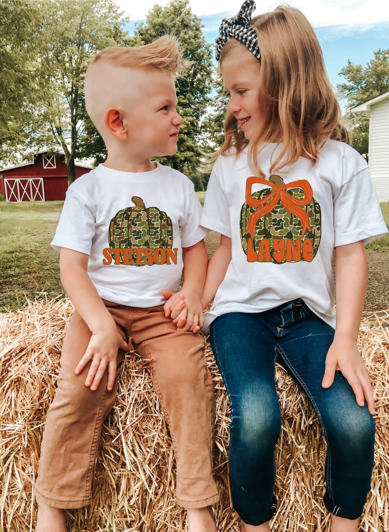 Two children wearing white t-shirts with custom pumpkin designs with names, sitting on a hay bale outdoors.