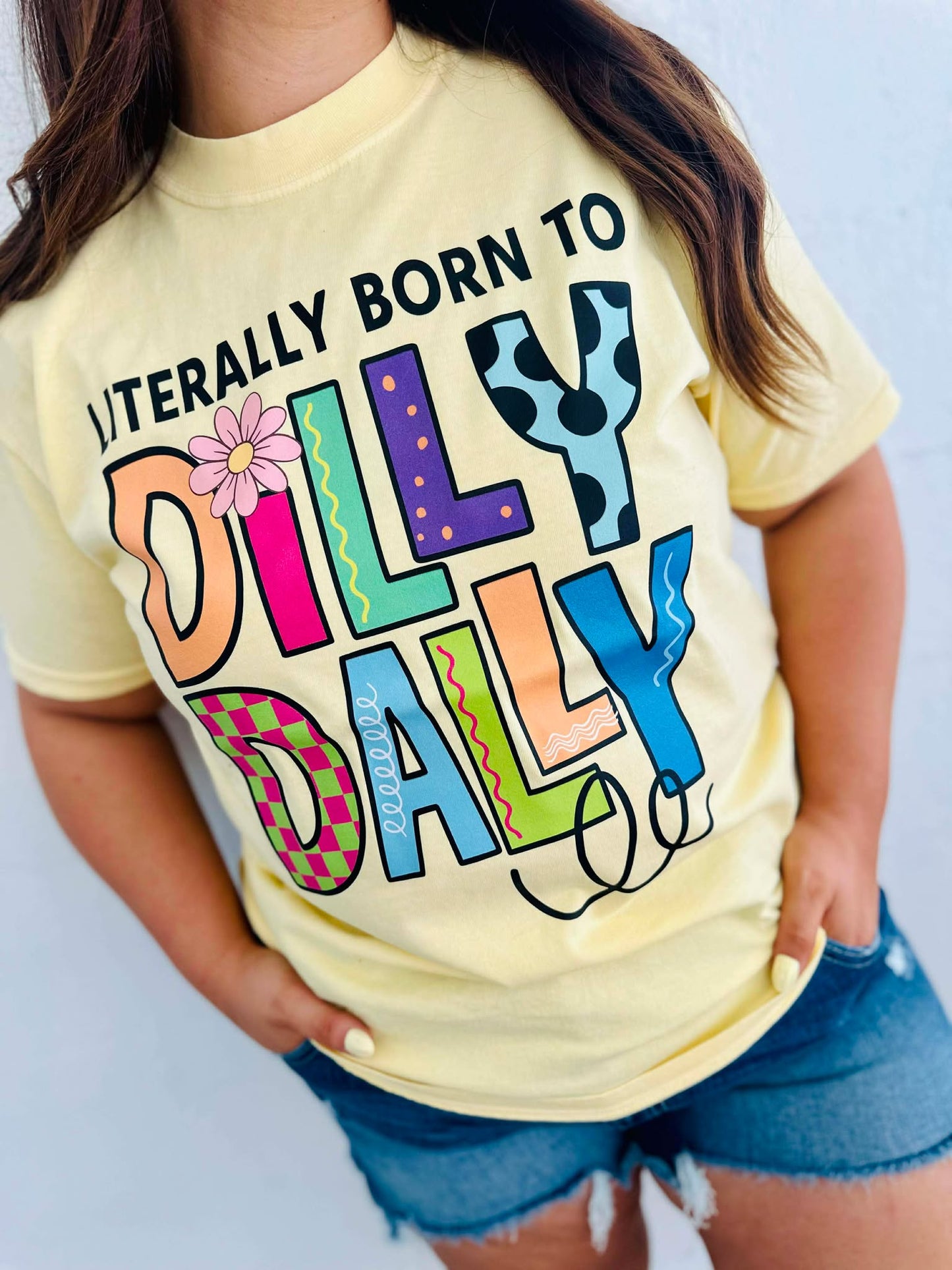 Person wearing a yellow t-shirt with colorful Born to Dilly Dally text design on a plain background