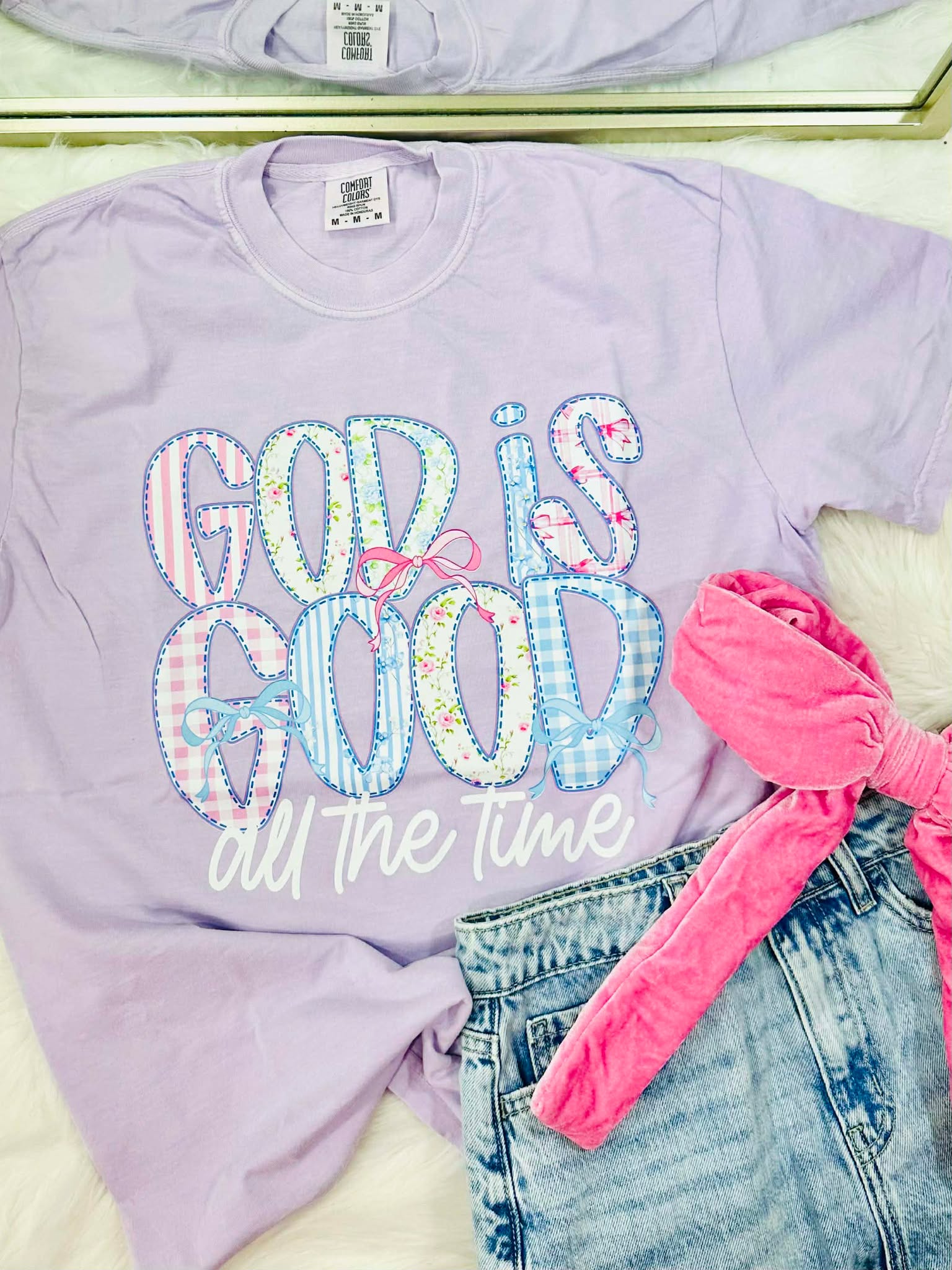 Lavender sweatshirt with 'God is good all the time' text, pink bow, and denim shorts.