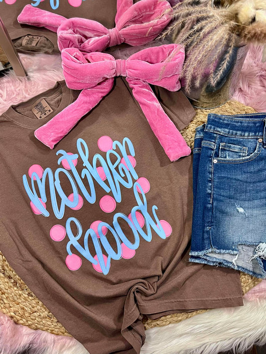 Brown t-shirt with 'Mother Hood' text and pink polka dots, paired with denim shorts and a pink bow on a textured surface.