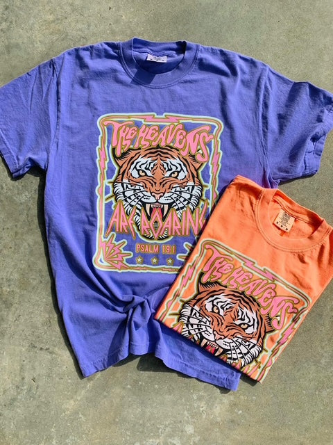 A graphic tee with a melon and violet color scheme featuring The Heavens Are Roaring Psalm 19:1 and a tiger design, laid flat on a surface.